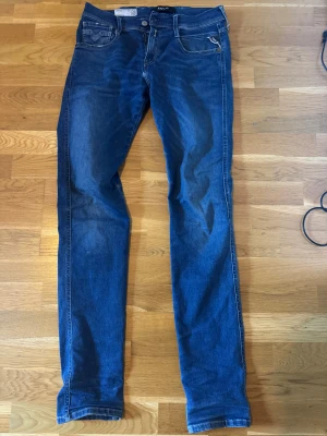 Blå Replay Anbass Hyperflex jeans - Säljer ett par blå Replay Anbass Hyperflex jeans med klassisk femficksdesign och snygga kontrastsömmar. Jeansen har lite skinny passform och är tillverkade i stretchigt denim för extra komfort. Märkespatch i skinn baktill och dragkedjegylf. De har lite slitningar men inget som syns tydligt eller förstör. 