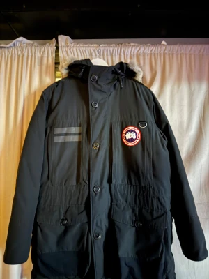 Svart Canada Goose parkas XL - Svart parkas från Canada Goose med ikonisk röd och vit patch på bröstet, huva med äkta päls och flera fickor framtill. Jackan har både dragkedja och knappar, samt snörning vid huvan. Perfekt för kalla vinterdagar och har en loose passform. Finns kvitto bevis om det behövs 