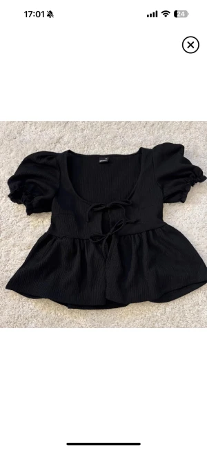 Svart blus med puffärm från Gina Tricot - Supersöt svart blus från Gina Tricot i storlek XS. Blusen har puffiga korta ärmar, v-ringning och två knytband framtill. Materialet är ribbat och ger en snygg struktur. Passformen är loose och den har en lätt volang nedtill.