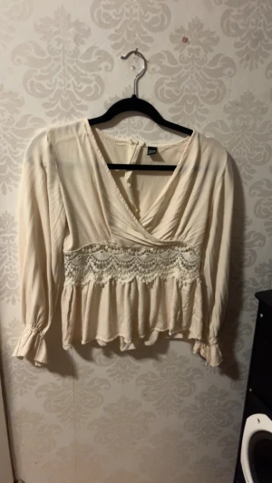 Beige blus med spets och volang - Supersöt beige blus från Lindex med omlott-v-ringning, spetsdetalj i midjan och volang nertill. Långa ärmar med volang vid ärmslut ger en romantisk vibe. Perfekt att styla med jeans eller kjol för en trendig look.