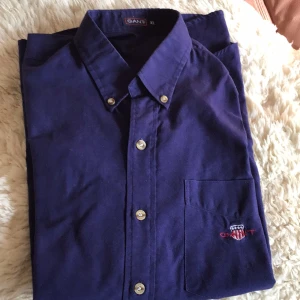 Mörkblå skjorta från GANT - Säljer en stilren mörkblå skjorta från GANT med klassisk krage och knappar framtill. Skjortan har en bröstficka med broderad logotyp. Perfekt för en snygg och avslappnad look.