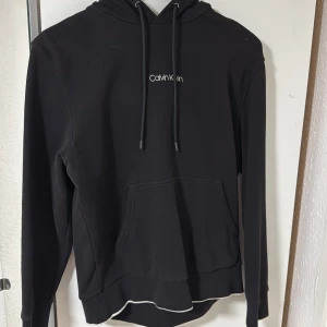 Svart hoodie från Calvin Klein - Svart hoodie från Calvin Klein med vit logga på bröstet. Tröjan har huva med snörning, känguruficka och ribbade muddar. Mjuk bomullskänsla och klassisk passform, perfekt för chill dagar. Snygg och enkel design som funkar till allt. Storlek M