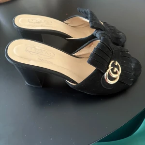 Svarta mocka klackpumps med tofsar - Svarta pumps i mocka med bred klack och öppen häl. Framsidan har dekorativa tofsar och en snygg guldfärgad GG-detalj. Skorna har en klassisk, rund tå och en lyxig känsla som lyfter varje outfit.