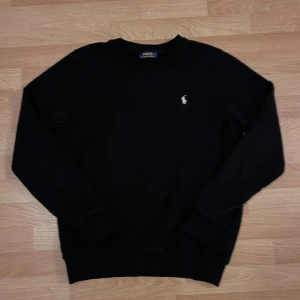 Svart tröja från Polo Ralph Lauren - Svart långärmad tröja från Polo Ralph Lauren med klassisk rund hals och broderad vit logga på bröstet. Tröjan har en enkel och stilren design med ribbade muddar vid ärmslut och nederkant. Perfekt för dig som gillar clean och tidlös stil.