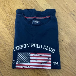 Marinblå t-shirt Vinson Polo Club XL - Marinblå t-shirt från Vinson Polo Club i bomull. Framsidan har tryck med amerikansk flagga och texten 'Vinson Polo Club'. Klassisk rund hals och korta ärmar. Snygga röda och vita detaljer vid halsringningen och trycket.