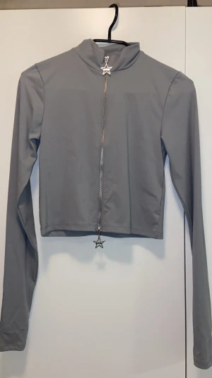 Grå croppad polotopp från FB Sister - Snygg grå croppad polotopp från FB Sister med lång ärm och cool silverfärgad dragkedja framtill. Dragkedjan har en stjärnformad detalj med FB-logga. Toppens material känns mjukt och stretchigt, perfekt för dig som gillar stilrena och moderna plagg.