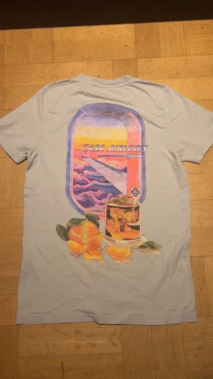 Casa Airlines t-shirt från Casablanca - Snygg ljusgrå t-shirt från Casablanca med färgglad Casa Airlines-tryck på både bröstet och ryggen. Motivet visar ett flygplansfönster med solnedgång, drink och apelsiner. Mjuk bomull och klassisk passform, perfekt för dig som gillar unika prints.