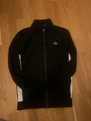 Svart ziptröja Lacoste Sport XS - Svart ziptröja från Lacoste Sport i klassisk passform, storlek XS. Tröjan har hel dragkedja, ståkrage och vita paneler längs sidorna. Ikoniska Lacoste-loggan på bröstet och stor logotyp på ryggen. Perfekt för en sportig och clean look.