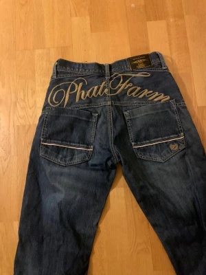 Phat Farm blå jeans med broderi - Säljer ett par blå jeans från Phat Farm med stort broderat logotyp på baksidan och coola detaljer på bakfickorna. Jeansen har en rak passform och klassisk femficksdesign. Materialet är slitstarkt denim och färgen är mörkblå med lätt tvättade partier.