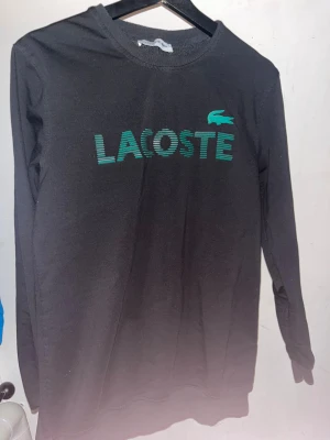 Svart långärmad Lacoste tröja - Svart långärmad tröja från Lacoste med grönt tryck på bröstet.