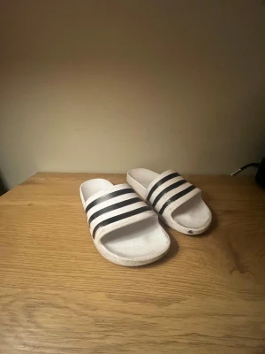 Adidas vita tofflor med svarta ränder - Säljer ett par klassiska Adidas sandaler i vitt med tre svarta ränder över foten. De har en platt sula och öppen tå, tillverkade i syntetmaterial. Perfekt för chill dagar och enkel att dra på sig. Snygg och sportig look med tydlig Adidas-logga under sulan.