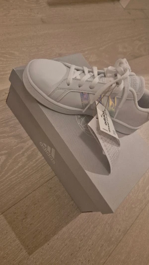 Adidas Grand Court K vita sneakers - Adidas Grand Court K sneakers i vitt med holografiska detaljer på sidan. Klassisk låg modell med snörning och rund tå. Snygg och stilren design med mjuk syntetöverdel och platt sula. Perfekt för dig som gillar cleana sneakers med en twist. Barnskor