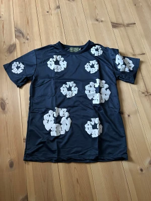 Blå t-shirt med vita blommor Denim Tears - Snygg mörkblå t-shirt från Denim Tears med stora vita blommönster över hela plagget. T-shirten har rund hals och korta ärmar, tillverkad i mjuk bomull som känns skön mot huden. Perfekt för dig som gillar streetwear och unika prints.