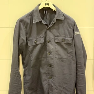 Mörkblå overshirt från Stone Island  - Snygg mörkblå overshirt från stone island med cool snörning i halsen och två bröstfickor med knappar. Skjortan har lång ärm och klassisk krage, perfekt för dig som gillar stilrena detaljer och funktionell design. Finns bara 74 stycken.