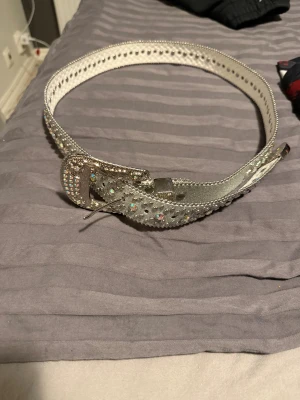 Glittrigt silverbälte med strass - Säljer ett snyggt silverfärgat bälte med glittrande strassdetaljer och dekorativa nitar längs hela kanten. Bältet har en bred, glittrig spänne och är gjort i syntetmaterial med en ljus insida. Perfekt för dig som vill sticka ut med lite extra bling.