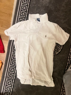 Vit piké från Polo Ralph Lauren - Vit pikétröja från Polo Ralph Lauren med klassisk krage, knappar framtill och den ikoniska broderade loggan på bröstet. Tröjan är i bomull och har korta ärmar, perfekt för en clean och stilren look.