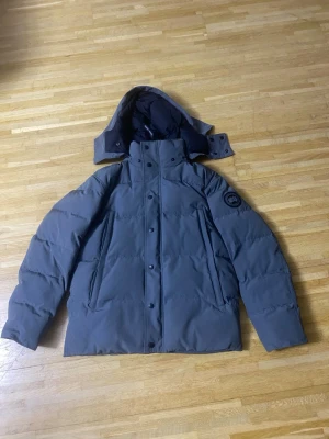 Canada gosse jacka i storlek M - Säljer en mörkgrå dunjacka från Canada Goose med avtagbar huva och klassisk logga på ärmen. Jackan är quiltad, har dragkedja och knappar framtill samt två stora fickor. Perfekt för kalla vinterdagar och riktigt snygg streetstil.