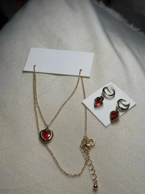 Hjärtformat Smyckeset – Halsband & Örhängen - Fint smyckeset i guldton med röda hjärtan. Innehåller halsband och matchande örhängen – båda helt nya. Perfekt present eller julklapp.  💰 Nypris: ca 99–149 kr per del (värde ca 200–300 kr totalt). 