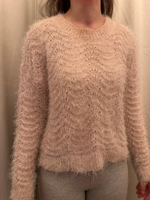 Ljusrosa  fluffig stickad tröja - Supermysig ljusrosa stickad tröja med fluffig yta och vågigt hålmönster. Tröjan har rund hals och långa ärmar, perfekt för dig som gillar en cozy vibe. Passformen är normal och materialet känns mjukt mot huden.