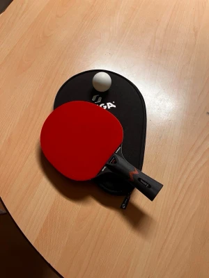 Röd och svart pingisracket STIGA - Säljer en STIGA pingisracket med ena sidan i intensivt rött och andra i svart. Handtaget är svart med röd detalj och texten 'Royal Carbon'. Racketen har en klassisk rund form och medföljer ett svart fodral med STIGA-logga samt en vit pingisboll.