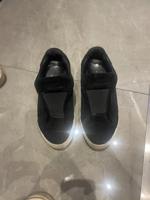 Svarta sneakers från Axel Arigato - Säljer ett par svarta slip-on sneakers från Axel Arigato med vit platt sula och rund tå. Ovandelen är i mocka och har en stilren design utan snörning. Perfekt för dig som gillar minimalistisk stil och vill ha något som funkar till jeans eller shorts. Dom är super fina på och säljer endast för att jag vill ha pengar för de istället!! 