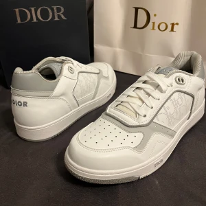 Vita Dior sneakers med grå detaljer - Snygga vita Dior sneakers med grå detaljer och diskret Dior-mönster på sidorna. Skorna har rund tå, platt sula och snörning framtill. Tillverkade i skinn med perforeringar på tån för extra ventilation. Ikonisk Dior-logga på hälen och tungan.