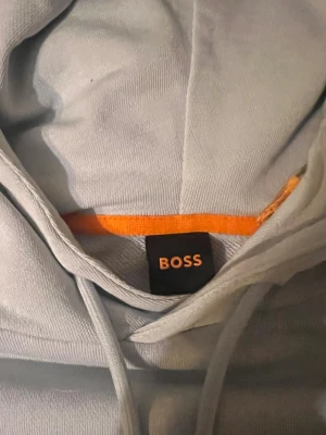 Babyblå färg  hoodie från BOSS - Snygg babyblå  hoodie från BOSS med broderad logga på bröstet. Tröjan har huva med snörning och en stor magficka framtill. Tillverkad i mjuk bomull som är skön att ha på sig. Perfekt för dig som gillar stilrena och cleana plagg.