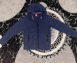 Marinblå Nike hoodie med dragkedja - Snygg marinblå hoodie från Nike med hel dragkedja framtill och två fickor med svarta dragkedjor. Tröjan har huva och långärmade ärmar, perfekt för chill eller träning. Materialet är mjukt och känns som bomull och polyester. Nike-logga i silver på bröstet.