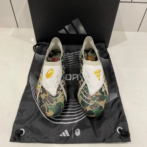 Adidas x BAPE fotbollsskor - Unika Adidas x BAPE fotbollsskor med grön, brun och beige kamouflagemönster, vita detaljer och guldiga inslag. Insidan har BAPE-typiska grafik och loggor. Perfekt för dig som vill sticka ut på planen. Man får såklart med original låda och påse. Hör gärna av er vid frågor!