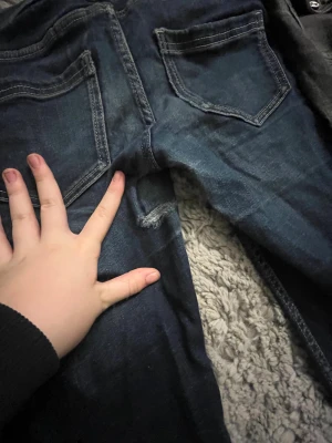 Bootcut jeans i blått och grått - Två par bootcut jeans, ett i mörkblå denim och ett i grått. Båda har klassiska femficksdetaljer och kontrastsömmar. De blå har dekorativa ficklock med knapp framtill och markerade bakfickor. Materialet är jeans och passformen är normal med utsvängda ben.