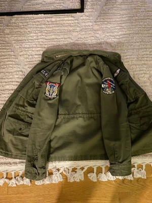 Olivgrön parkajacka Polo Ralph Lauren - Säljer en olivgrön parkajacka från Polo Ralph Lauren med coola militärinspirerade patches på ärmarna. Jackan har dragkedja, flera fickor och klassisk parkamodell. Perfekt för dig som gillar utility-stil och streetwear.