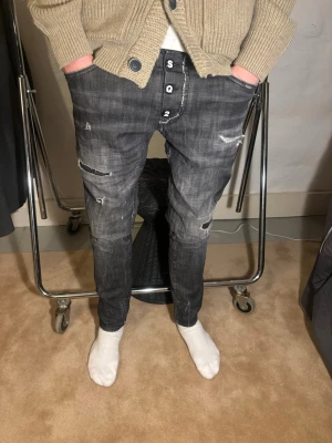 Dsquared2 jeans - Säljer ett par grå jeans från Dsquared2, modell 1964. De har en snygg sliten look med flera slitningar och detaljerade knappar framtill. Jeansen har raka ben och klassiska bakfickor, tillverkade i jeansmaterial med en cool tvättad finish. Modellen på bilden är 179 cm lång. Storlek 46, motsvarar 31. Nytt pris 6000kr mitt pris 349kr✅Vid funderingar hör av dig 📩