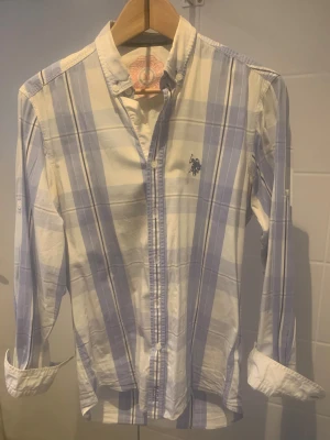 Rutig skjorta från US Polo Assn - Snygg rutig skjorta från US Polo Assn i vitt och ljusblått. Klassisk button-down krage, broderad logga på bröstet och långa ärmar. Skjortan är i bomull och har knappar framtill. Perfekt för dig som gillar en clean och stilren look.