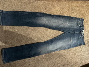 Replay Anbass jeans blå slim fit - Snygga Replay Anbass jeans i klassisk blå tvätt med slitningar och lätt distressed detaljer. Slim fit-modell med fem fickor, läderpatch bak och dragkedja i gylfen. Perfekt för dig som gillar en modern och stilren look.