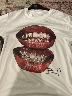 Vit t-shirt med grafiskt tryck från H&M - Snygg vit t-shirt från H&M med ett stort grafiskt tryck av två läppar med grillz och texten 'Be Real' och 'No Cap'. Trycket har coola detaljer och t-shirten har en klassisk rund hals. Perfekt för dig som gillar streetstyle och statement-plagg.