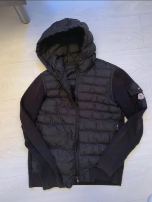 Svart dunjacka från Moncler med huva - Svart dunjacka från Moncler med quiltad front, stickade ärmar och stor huva. Jackan har dragkedja framtill och en Moncler-logga på ärmen. Perfekt för dig som vill ha en snygg och varm jacka med streetkänsla och coola detaljer.