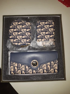 Blå och beige plånbok set RK - Snyggt set med plånbok, korthållare och myntfodral från RK. Beige och blått mönster med RK-logga och glittrig detalj framtill. Plånboken har lock med knapp och alla delar har ett coolt grafiskt tryck. Perfekt för att hålla koll på kort och cash.