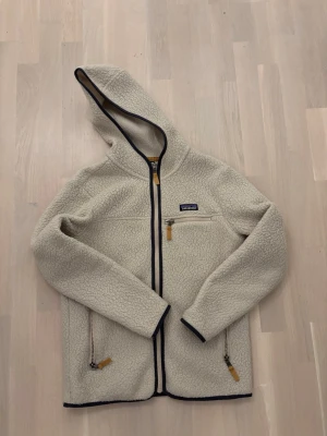 Patagonia fleece - Mysig beige teddyjacka från Patagonia med huva och svarta detaljer. Jackan har tre fickor med dragkedja och snygga bruna zip-pullers. Perfekt för kalla dagar, med långärmad design och mjukt fleece-material som håller dig varm.