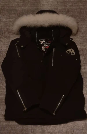 Svart dunjacka Moose Knuckles med päls - Svart dunjacka från Moose Knuckles med stor vit päls på huvan, silverfärgade dragkedjor och broderad patch på insidan. Jackan har flera fickor med dragkedja och klassisk logga på ärmen. Perfekt för kalla dagar och riktigt snygg streetstil. Köpt på Johnells förra året. 