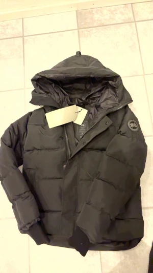 Svart pufferjacka från Canada Goose - Svart pufferjacka från Canada Goose med huva och diskret logotyp på ärmen. Jackan har quiltad design, dragkedja och muddar vid ärmslut. Perfekt för kalla dagar och har en clean, modern look som funkar till streetwear. Jackan är ganska tajt och passar M och L
