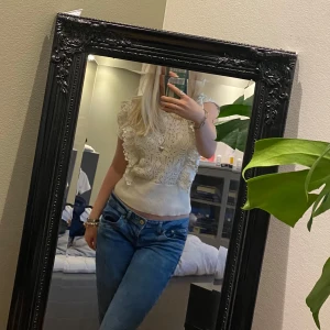 Vit spetsig blus med volangdetaljer - Supersöt vit blus med spetsmönster och volangdetaljer längs axlarna. Ärmlös modell med ribbad midja som ger en snygg passform. Perfekt för dig som gillar romantisk och feminin stil. Lätt att matcha med både jeans och kjol.💞