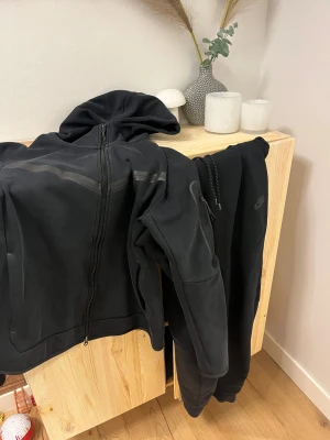Svart Nike hoodie + byxa - Svart hoodie och byxor från Nike med huva, dragkedja och snygga detaljer som snörning och ficka med dragkedja på ärmen. Tröjan har en stilren look med diskret Nike-logga på bröstet och är tillverkad i mjukt material som passar perfekt för chill eller träning.