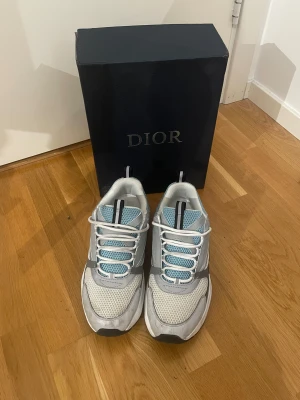 Dior B22 sneakers  - Säljer ett par Dior B22 sneakers med chunky silhuett i grått, vitt och blått. Skorna har meshpaneler, skinn och syntetdetaljer, samt vita snören och svart sula med blå inslag. Märket syns på sidan och tungan. Perfekt för dig som gillar exklusiva sneakers med streetkänsla. Storlek 45, bra skick, box medföljer