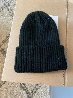 Svart ribbad mössa - Svart ribbstickad mössa i 100% akryl. Klassisk design med bred uppvikt kant och snygg struktur som passar till alla jackor. Perfekt för kalla dagar och enkel att matcha med din stil.