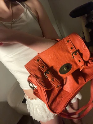 Orange y2k vintage axelväska i skinn med spännen - Snygg orange axelväska i skinn med coola metallspännen och ett vridlås framtill. Väskan har både handtag och lång axelrem, så du kan bära den på olika sätt. Flera detaljer i antikmässing och praktiska fack gör den både stilren och användbar. Perfekt för dig som gillar färg och vill sticka ut. Metall delar kan ha ändrat färg eller rostat då den är vintage.