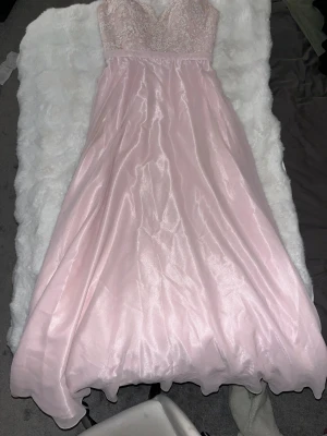 Ljusrosa långklänning med spetsliv - Drömmig ljusrosa långklänning från Ivory Fields med glansigt tyg och vackert spetsbroderat liv. Klänningen har v-ringning, smala axelband och snörning i ryggen för perfekt passform. Kjolen är svepande och ger en elegant siluett, perfekt för speciella tillfällen. Går att justera till behov. Superfin klänning till bal samt övriga tillfällen. Endast använd i en kväll, köpt för 4000!