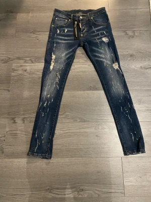 Dsquared2 skinny jeans med dragkedja - Mörkblå skinny jeans från Dsquared2 med slitningar, färgstänk och en cool dragkedjedetalj framtill. Klassisk femficksmodell med gul kontrastsöm och Dsquared2-logga vid gylfen. Perfekt för dig som gillar streetstyle och edgy detaljer.