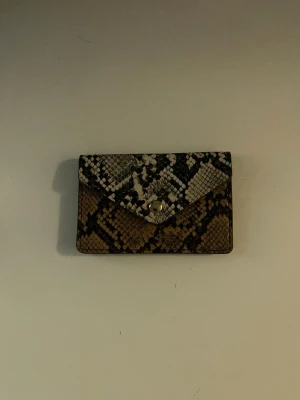 Plånbok i ormskinn - Snygg plånbok med ormskinnsmönster i läder. Plånboken har kuvertformad design med tryckknapp i guld och flera fack på insidan. Perfekt storlek för kort och sedlar. Har köpts på glitter för några år sedan men har inte använts.