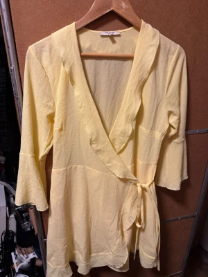 Gul omlottblus från Sanne Alexandra - Snygg gul omlottblus från Sanne Alexandra med trekvartsärm och knytband i midjan. Blusen har en lätt struktur i tyget och volangkant längs omlottdelen. Perfekt för dig som gillar ljusa färger och en feminin look.