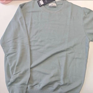 Ljusgrå sweatshirt från Stone Island - Snygg ljusgrå sweatshirt från Stone Island med klassisk rund hals och ribbade muddar vid ärmslut och nederkant. Perfekt för dig som gillar stilrena och enkla plagg med premiumkänsla. Taggen sitter kvar och materialet är mjuk bomull. Dm vid intresse